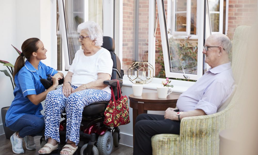 Male And Female Residents Sitting In Chair And Talking With Nurse In Retirement Home