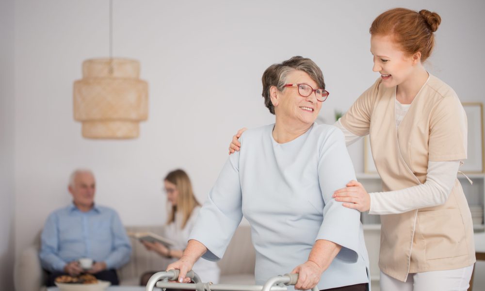 Happy nurse assisting a senior woman with a walker and another nurse reading a book to a pensioner in the background